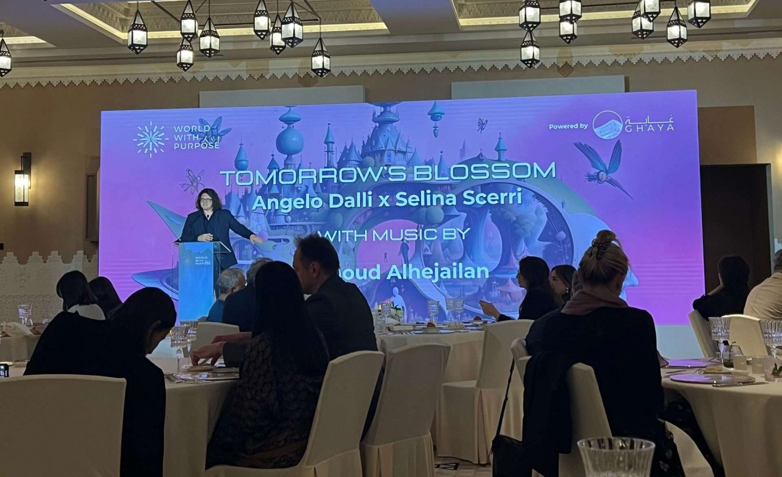 Angelo Dalli & Selina Scerri & CSAI institute brought ‘Tomorrow’s Blossoms’ to World With Purpose conference at the UNESCO world heritage site of Diriyah in Saudi Arabia. In the background is Angelo speaking with the Ai video played on the screen behind him whilst in the foreground people watch whilst seated at their tables.