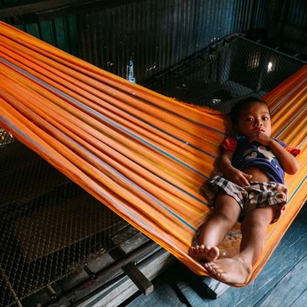 This photograph was taken by Jacob Sammut and it depicts a Cambodian boy seated on an orange hammock whilst looking at the photographer.