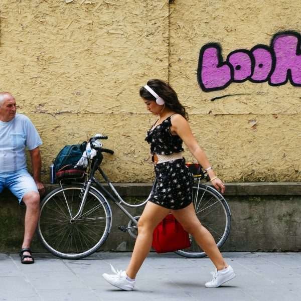 A coloured photograph showing a woman crossing from the right side of the photograph to the left whilst looking down. SHe wears a short black skirt with small designs and a matching top whilst she wears white jogging shoes and white headphones. On the yellow wall in the background is a bright pink graffiti with black outlines with the word 'look'. Behind her, resting against the wall, is a bike and in front of it, is a man dressed in a blue tshirt and blue shorts and black sandals seated on the wall to the left of the photograph The man is seen looking at the woman passing by. Above himis a surveillance camera signage.