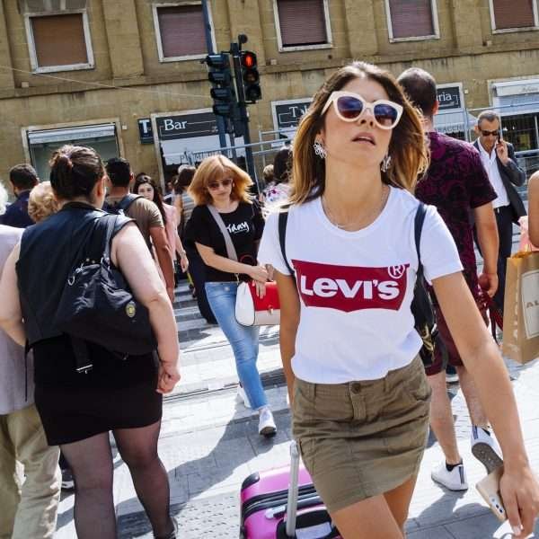 This is a coloured photograph by Jacob Sammut howing a white girl walking confidentally towards the photographer in a white tshirt and a red logo of 'Levis' across the front and a green skirt and white sunglasses. A crowd of people are seen walking across the road where traffic lights stand. The photograph was taken in Florence.