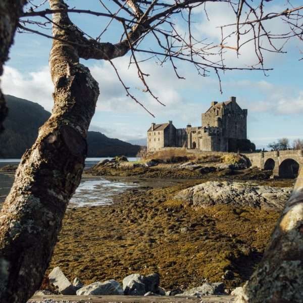Photograph of the Eilean Donan Castle in Scotland, taken by Jacob Sammut. In the foreground are trees and grass and rocks with water and the castle in the background. The sky has some visible clouds.